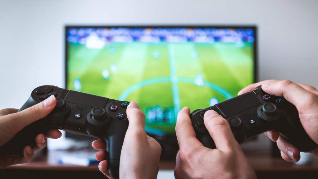 Dos personas con Mandos de Playstation  jugando un videojuego en una pantalla de fondo 